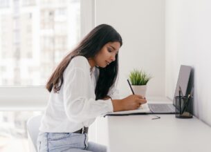 A student takes notes in front of her laptop on a white desk
