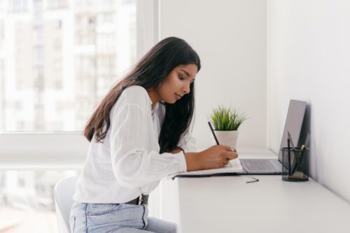 A student takes notes in front of her laptop on a white desk